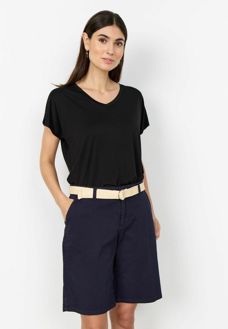 Femme portant un haut noir à col en V, un short bleu marine jusqu'aux genoux et une ceinture tressée beige, debout les mains dans les poches devant un fond uni.