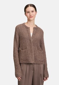 Frau, die einen braunen, flauschigen Cardigan mit zwei Fronttaschen über taubfarbenen, weit geschnittenen Hosen trägt und vor einem schlichten, hellen Hintergrund steht.