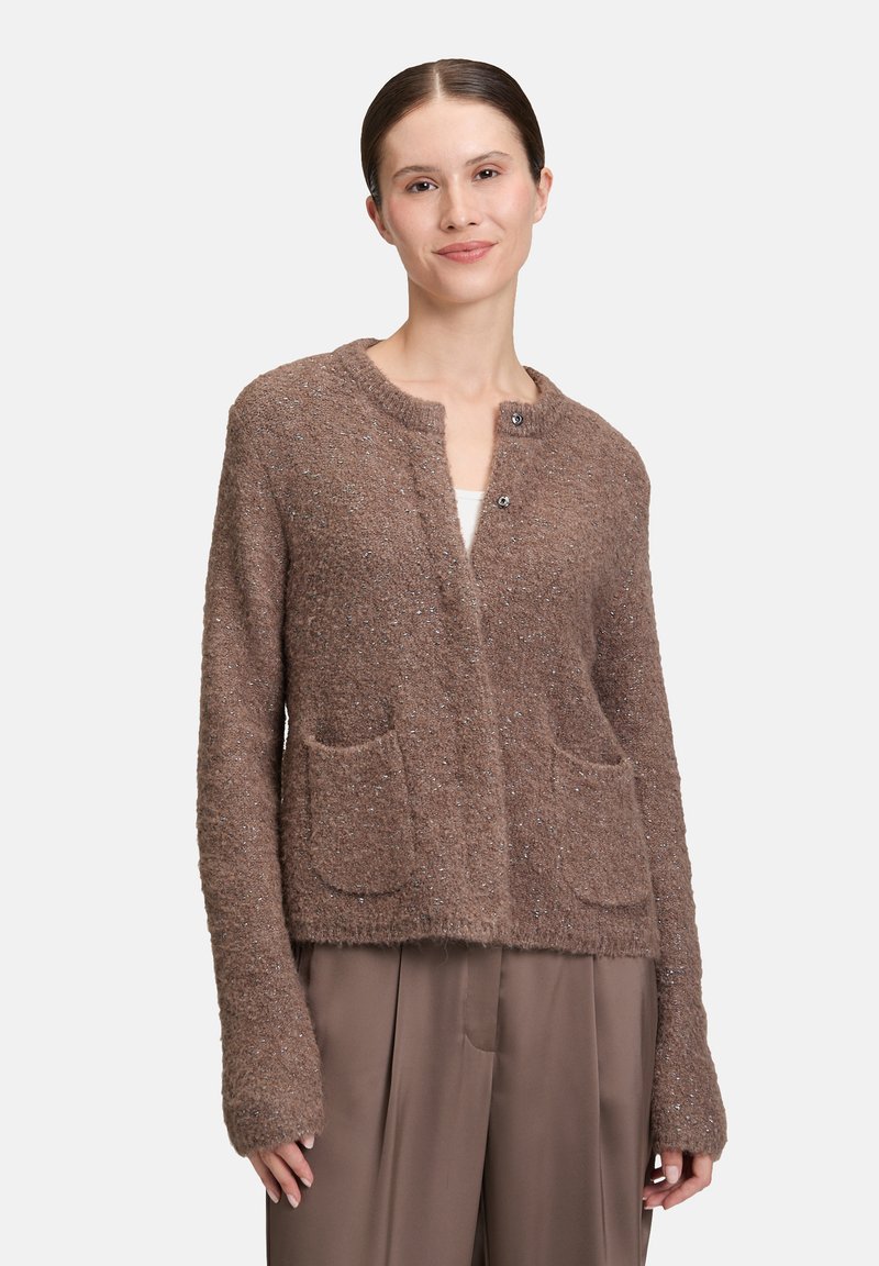 Frau, die einen braunen, flauschigen Cardigan mit zwei Fronttaschen über taubfarbenen, weit geschnittenen Hosen trägt und vor einem schlichten, hellen Hintergrund steht.