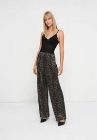 Femeie în picioare, îmbrăcată cu un top fără mâneci de culoare neagră, pantaloni largi cu imprimeu de leopard și pantofi cu vârf ascuțit negri, pe un fundal alb simplu.