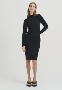Schwarzes langärmliges Kleid mit hohem Ausschnitt, figurbetonter Silhouette, strukturiertem Stoff und seitlichem Schlitz, kombiniert mit schwarzen Plateausandalen.