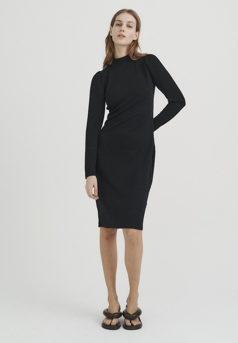 Schwarzes langärmliges Kleid mit hohem Ausschnitt, figurbetonter Silhouette, strukturiertem Stoff und seitlichem Schlitz, kombiniert mit schwarzen Plateausandalen.