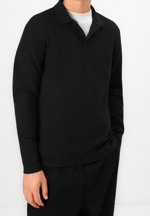 Homme portant un polo à manches longues noir sur un sous-vêtement blanc, associé à un pantalon noir, debout devant un fond uni.
