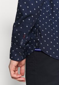 Camisa de color marino con un pequeño patrón espiral blanco, puños con botón y etiqueta visible en el lateral. La tela parece suave y ligera.