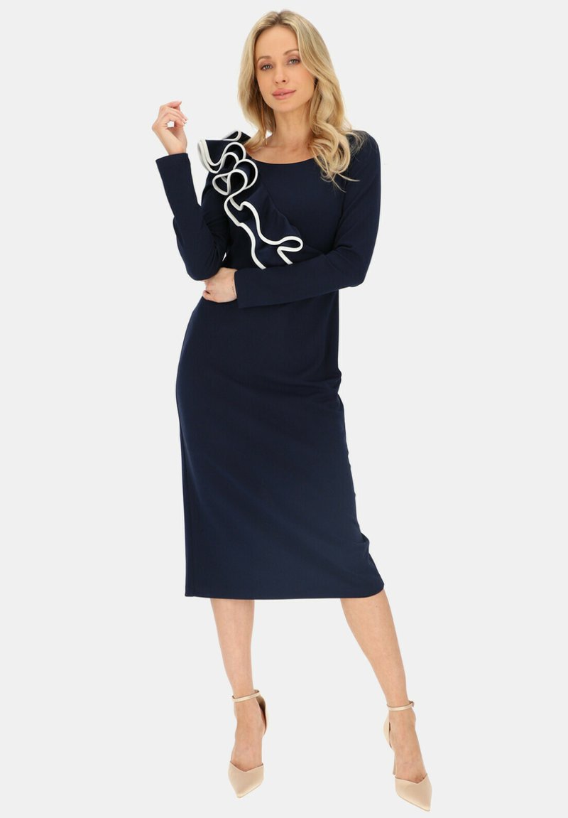 Granatowa sukienka z długim rękawem z falbankami w białym kolorze na lewym ramieniu, długości do kolan, wykonana z gładkiego materiału. Gładka faktura, dopasowany krój.