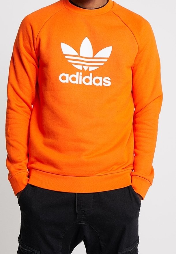 Oranžová fleecová mikina s kulatým výstřihem, dlouhými rukávy a žebrovanými manžetami. Na přední straně je velké bílé logo Adidas trefoil.