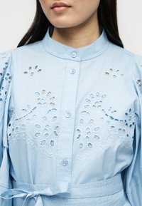 Γυναίκα που φοράει γαλάζιο πουκάμισο με κουμπιά, διακοσμημένο με κεντήματα λουλουδιών σε αποκοπή και ζώνη στη μέση, εμφανίζεται από τους ώμους έως τη μέση.