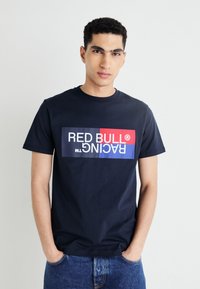 Tamsiai mėlynas medvilninis marškinėlis su ryškiu grafikiniu „RED BULL RACING“ logotipu baltos, raudonos ir mėlynos spalvų derinyje, esantis priekyje. Trumpomis rankovėmis, apvaliu kaklu.