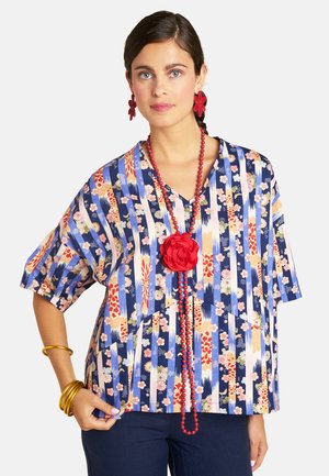 Woman with dark hair wearing a blue floral striped blouse, red beaded necklace with large flower, red flower earrings, and gold bangles.