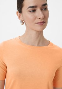 Koszulka z krótkim rękawem i okrągłym dekoltem w miękkim koralowym kolorze. Posiada gładką teksturę i minimalistyczny design, w połączeniu z srebrnymi kolczykami w kształcie kropli.