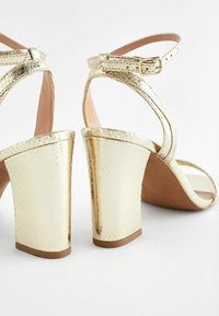 Guld hælhøje sandaler med en tekstureret metallisk finish, ankelrem og blokhælsdesign. Åben tå med en glat inderside.