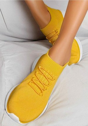 Legs wearing bright yellow knit slip-on sneakers with laces, resting on a white cushioned surface.