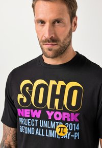 Černé tričko s výrazným vícebarevným textem: "SOHO," "NEW YORK," a dalšími grafikami v růžové, modré a žluté. Krátké rukávy, kulatý výstřih.
