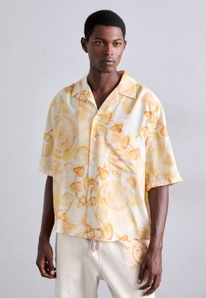Chemise à manches courtes de couleur jaune pâle avec un motif tropical et du texte. Dotée d'une poche poitrine et d'une coupe décontractée. Portée avec un pantalon de couleur claire.