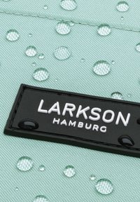 Zöld texturált szövet vízcseppekkel, amelyen egy fekete téglalap alakú címke található, amelyen fehér betűkkel az "LARKSON HAMBURG" felirat olvasható.
