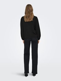 Svart texturerad blus med puffärmar, matchad med mörka jeans med raka ben och svarta klackar, sedd bakifrån. Mjukt hår faller ner.