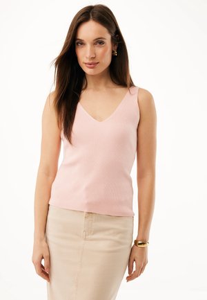 Kobieta o długich brązowych włosach, nosząca złote kolczyki-koła, różowy prążkowany top bez rękawów, beżową spódnicę oraz złotą bransoletkę, stojąca na białym tle.