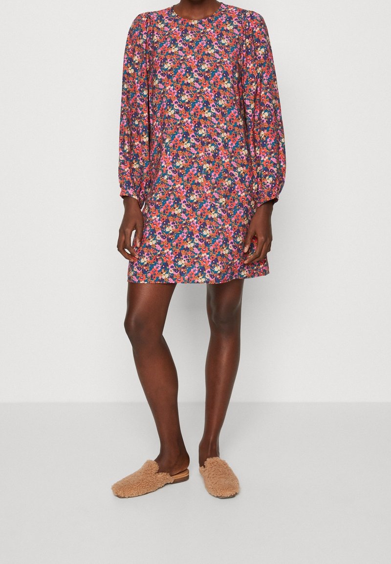 Femme portant une mini-robe à manches longues à fleurs multicolores et des chaussures beige pelucheuses sans lacets, debout devant un fond blanc uni.
