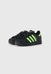 Baskets Adidas Superstar noires avec des rayures vert néon, un onglet au talon et un marquage, présentées côte à côte sur un fond blanc.