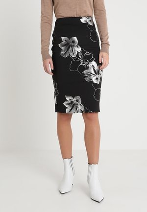 Femme portant une jupe crayon noire avec de grandes impressions florales blanches, un haut beige à manches longues et des bottines blanches, debout sur un fond uni.