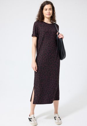 Frau trägt ein dunkelviolettes und schwarzes Midikleid mit Leopardenmuster und seitlichen Schlitzen, weiße Sneakers und trägt eine schwarze Umhängetasche.