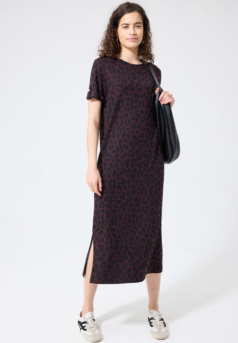 Femme portant une robe midi imprimé léopard violet foncé et noir avec des fentes sur les côtés, des baskets blanches, et un sac à bandoulière noir.