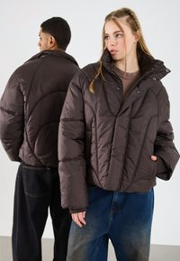 Două modele purtând jachete puffer de culoare maro închis; femeia cu fața către spectator, având mâna în buzunar, iar bărbatul cu fața lateral, amândoi purtând pantaloni casual din denim.