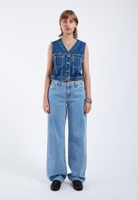 Chaleco de mezclilla con escote en V y botones, combinado con jeans de pierna amplia en azul claro. Ambos artículos tienen una textura desgastada y un ajuste casual.
