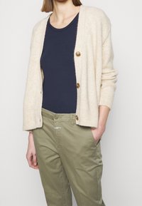 Beige, mjuk cardigan med avslappnad passform, tre stora bruna knappar och ribbade ärmslut. Bärs över en marinblå skjorta och matchas med olivgröna byxor.