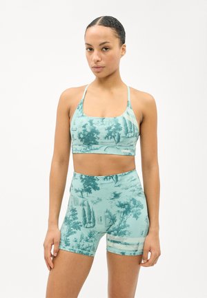 Femeie stând cu fața înainte, purtând un sutien sportiv cu model teal și pantaloni scurți de ciclism cu talie înaltă, asortați, cu dungi din plasă pe coapsă.