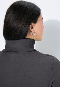 Una donna con capelli lunghi e lisci di colore scuro indossa orecchini a cerchio d'argento e un maglione a dolcevita grigio scuro a coste, voltata di spalle su uno sfondo bianco.