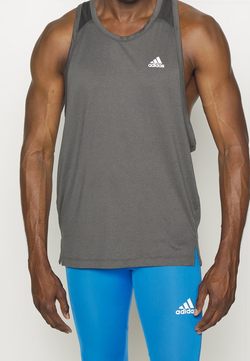Muž v šedém bez rukávů tričku Adidas a jasně modrých legínách Adidas, stojící na jednobarevném světlém pozadí.