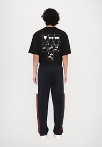 Sort t-shirt med et grafisk tryk af spillekort med hjerter på ryggen; parret med marineblå bukser med farveblokering på siden.