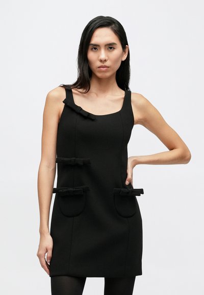 Fekete ujjatlan ruha négyzet alakú nyakkivágással, a zsebeknél három masnival díszítve, sima textúrával. Testhez simuló sziluett, minimális varrással.