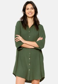 Short green button-up dress with short sleeves, featuring a slightly curved hem and a smooth texture. Gold buttons add a subtle accent.