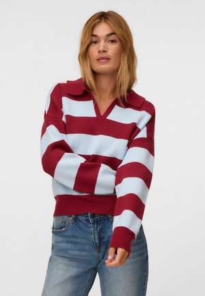 Jeune femme portant un pull large à rayures rouges et bleu pâle avec un col, et un jean bleu, sur un fond clair uni.