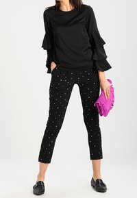 Zwarte top met gelaagde ruffle-mouwen, gecombineerd met zwarte broek met kleine witte hartpatronen, en een paarse clutch in de hand.