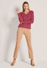 Eine Frau steht gegen eine schlichte Wand, trägt einen rosa Strickpulli, beige Slim-Pants und braune Absatzsandalen, und posiert mit einem erhobenen Arm.