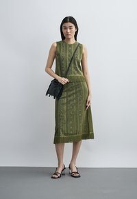Femme portant une robe sans manches vert olive à motifs avec des franges, un sac bandoulière noir et des talons ouverts noirs, debout contre un mur uni.