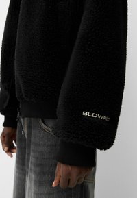 Svart, fluffig fleecejacka med ribbade ärmslut och midja. Har en dragkedjestängning och broderad "BLDWRLD"-logotyp på ärmen.