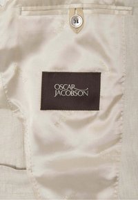 Beige textilní kapsa s tmavohnědým obdélníkovým štítkem, na kterém je bílý text "OSCAR JACOBSON." Zahrnuje detail kovového knoflíku. Měkká struktura.