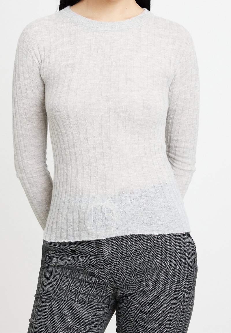 Donna che indossa un maglione aderente beige a coste con maniche lunghe e pantaloni grigi a motivo herringbone, in piedi davanti a uno sfondo semplice.