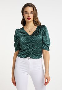 Grøn satinsbluse med V-hals, draperinger foran, pufærmer og et geometrisk mønster, parret med hvide jeans.