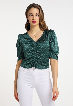 Blusa de satén verde con escote en V, fruncido en la parte delantera, mangas abullonadas y un patrón geométrico, combinada con jeans blancos.
