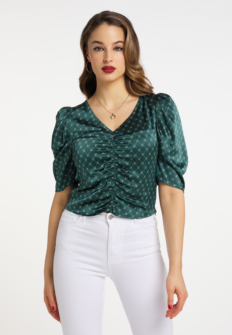 Grøn satinsbluse med V-hals, draperinger foran, pufærmer og et geometrisk mønster, parret med hvide jeans.