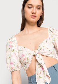 Top floral curto com mangas bufantes, apresentando um laço na frente. O tecido tem padrões florais em rosa e verde sobre um fundo claro.