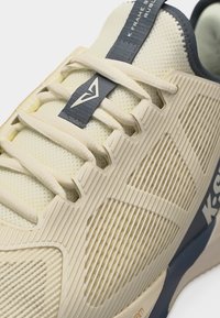 Zbliżenie na beżowo-granatowy but sportowy z teksturowaną syntetyczną cholewką, kremowymi sznurówkami oraz logo marki na języku i boku.