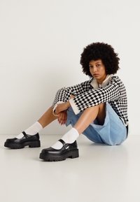 Maglione a scacchi bianco e nero, gonna in denim azzurro chiaro, calze bianche e scarpe con piattaforma nera e dettagli con cinturino, in posa seduta.