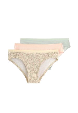 Nainen seisoo paljain jaloin yksiväristä taustaa vasten, yllään valkoinen pitsinen bralette ja siihen sopivat valkoiset alushousut.