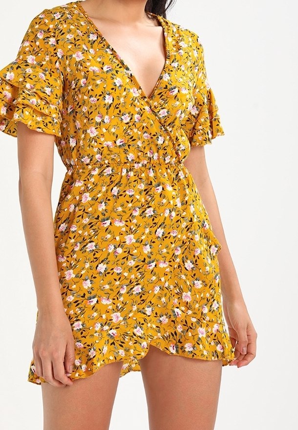 Gul blommig omlottklänning med korta volangärmar, V-ringning och elastisk midja. Har rosa och svarta blommönster på ett texturerat tyg.